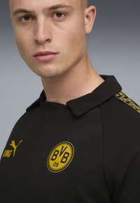 Sort sweatshirt med krave, der har et gult "BVB 09" logo og gult Puma-branding på skulderen. Glat tekstur, afslappet design.