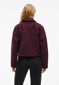 Veste en polaire bourgogne au design court, col à revers et épaules tombantes ; tissu texturé ; associée à un pantalon noir.