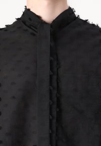 Black shirt with a high neckline, featuring a textured fabric adorned with small tufted accents throughout the material.