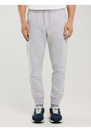 Muž nosící světle šedé tepláky s elastickými manžetami a modré tenisky, stojící před jednoduchým světlým pozadím.