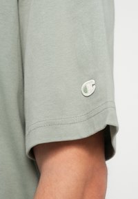 Tricou scurt din bumbac moale, de culoare verde deschis, cu mânecă scurtă, având un mic logo brodat în alb și verde pe mânecă.