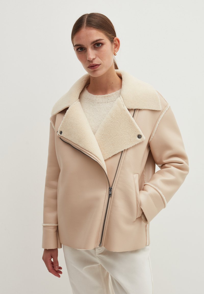 Frau in einer beigen Schaffelljacke mit großem Kragen, Reißverschlussdetails, cremefarbener Strickjacke und weißen Hosen, die vor einem schlichten weißen Hintergrund steht.