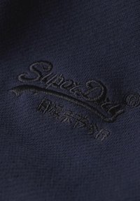 Tessuto blu navy con logo "Superdry" ricamato e design a corona. La texture appare liscia e i punti di cucitura sono dettagliati.