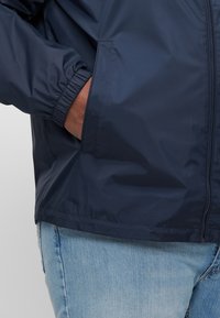 Mørkeblå lett jakke med elastiske mansjetter, lommer foran og glidelås. Glatt tekstur med et enkelt, funksjonelt design.