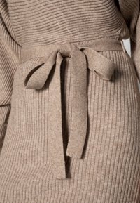Pull côtelé beige avec une texture douce, doté d'une ceinture nouée à la taille, mettant en valeur la silhouette.