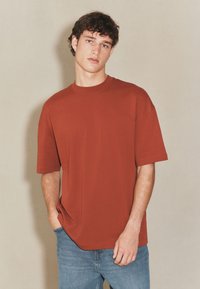 Przymarznięta, dużych rozmiarów koszulka w kolorze rdzy, wykonana z miękkiej bawełny, z okrągłym dekoltem i krótkimi rękawami. W zestawie z jasnoniebieskimi dżinsami.