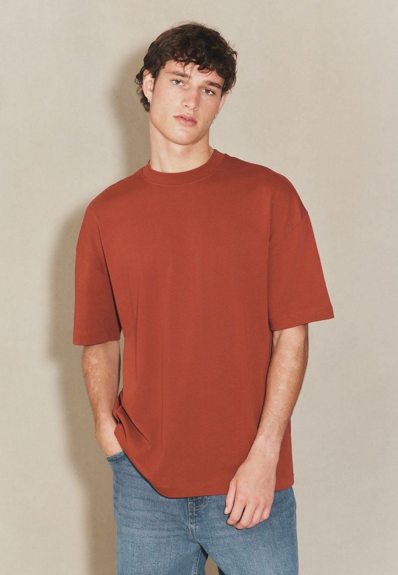 Przymarznięta, dużych rozmiarów koszulka w kolorze rdzy, wykonana z miękkiej bawełny, z okrągłym dekoltem i krótkimi rękawami. W zestawie z jasnoniebieskimi dżinsami.