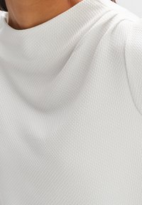 Top bianco realizzato in un tessuto testurizzato con un delicato motivo a griglia, colletto alto e morbide drappeggiature intorno alle spalle.