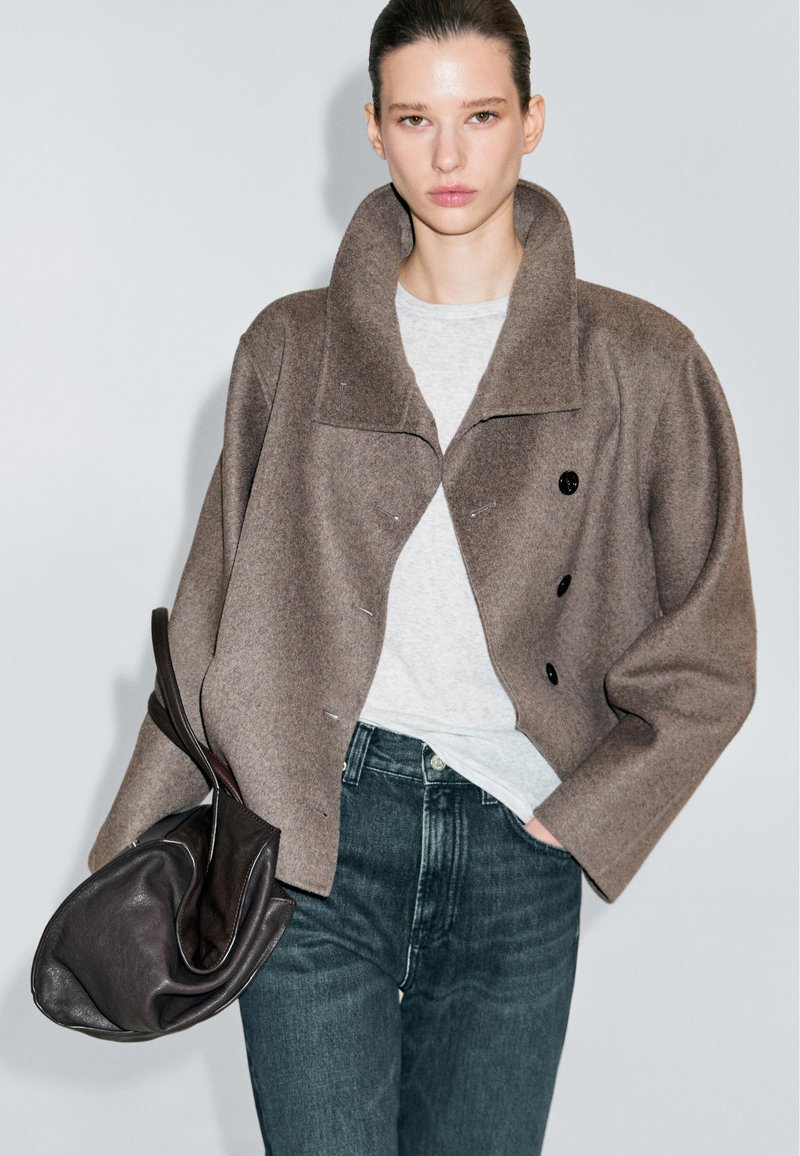 Donna che indossa un ampio giubbotto di lana marrone, una camicia grigio chiaro, jeans scuri, mentre tiene una grande borsa in pelle morbida con una mano in tasca.