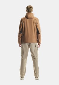 Hombre rubio de espaldas, vistiendo una chaqueta con capucha marrón y parches negros en los codos, pantalones cargo beige y zapatillas blancas, sobre un fondo blanco.