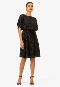 Robe noire avec des manches flutter courtes, des embellissements floraux en sequins, une taille cintrée et une jupe fluide. Associée à des talons à bride de cheville noirs.
