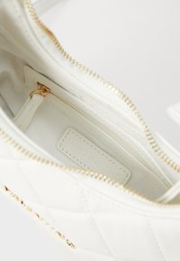 Wei�ß gefütterte Handtasche mit goldener Reißverschlus, ausgestattet mit einem Markenlabel. Sanfte Textur und dezente Fütterung sind sichtbar.