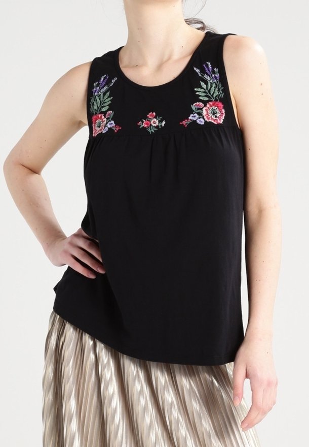 Femme portant un haut noir sans manches avec une broderie florale colorée au niveau de l'encolure, associé à une jupe plissée métallique.