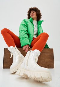 Person wearing bright green puffer jacket, orange leggings, and chunky white boots sitting on a wooden box against a plain background.