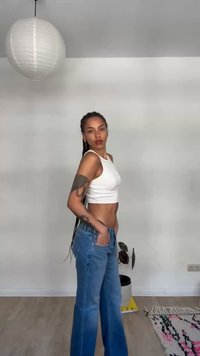 Γυναίκα με πλεξούδες και τατουάζ ποζάρει με λευκό crop top και μπλε τζιν σε ένα μινιμαλιστικό δωμάτιο με κρεμαστό φωτιστικό από χαρτί και χαλί.