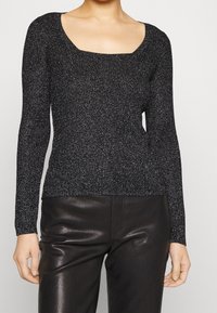 Nainen, joka pitää yllään mustaa ja hopeahippuista ribbikuvioista neulepuseroa, jossa on neliskulmainen pääntie, ja mustia nahkahousuja tasaisessa taustassa.