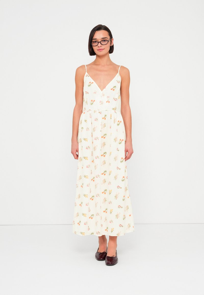 Μάξι φόρεμα με κρεμ φόντο, φλοράλ μοτίβο σε ροζ και πορτοκαλί, V-λαιμόκοψη, λεπτές τιράντες και ζώνη με πιέτες στη μέση· φορεμένο με σκούρα παπούτσια.