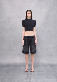 Camiseta negra corta hecha de un tejido suave, combinada con pantalones cortos de carga oscuros con bolsillos frontales y un lazo. Lleva puestas chanclas negras.