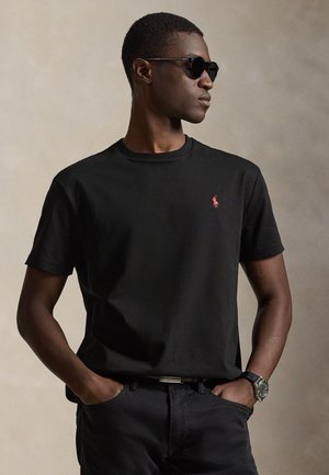 Homme portant un t-shirt noir avec un logo rouge, un pantalon noir, des lunettes de soleil et une montre-bracelet, debout les mains dans les poches devant un fond uni.