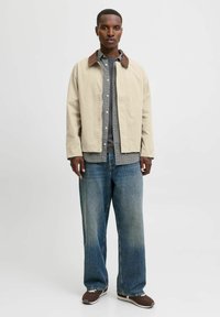 Beige jakke med brun krave over en ternet skjorte, parret med brede blå jeans og brune sneakers. Enkelt, praktisk design.