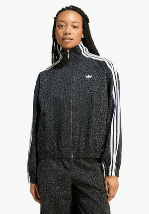 Donna che indossa una tuta Adidas nera con strisce bianche e un motivo geometrico sottile, in piedi davanti a uno sfondo chiaro e semplice.