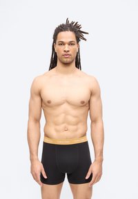 ing nélküli férfi dreadlock hajjal, fekete Calvin Klein boxeralsót visel, amelynek derékrésze barna színű, és előre néz.