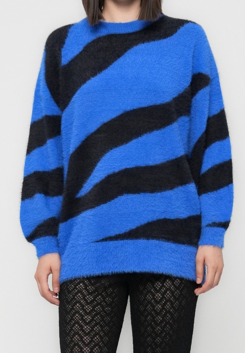 Pull bleu duveteux avec de larges rayures diagonales noires, porté sur un pantalon en dentelle noire, montré sur une personne des épaules jusqu'aux cuisses.