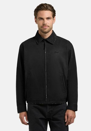 Mann med kort hår som bærer en svart jakke med glidelås og svarte bukser, stående mot en ensfarget lys bakgrunn.