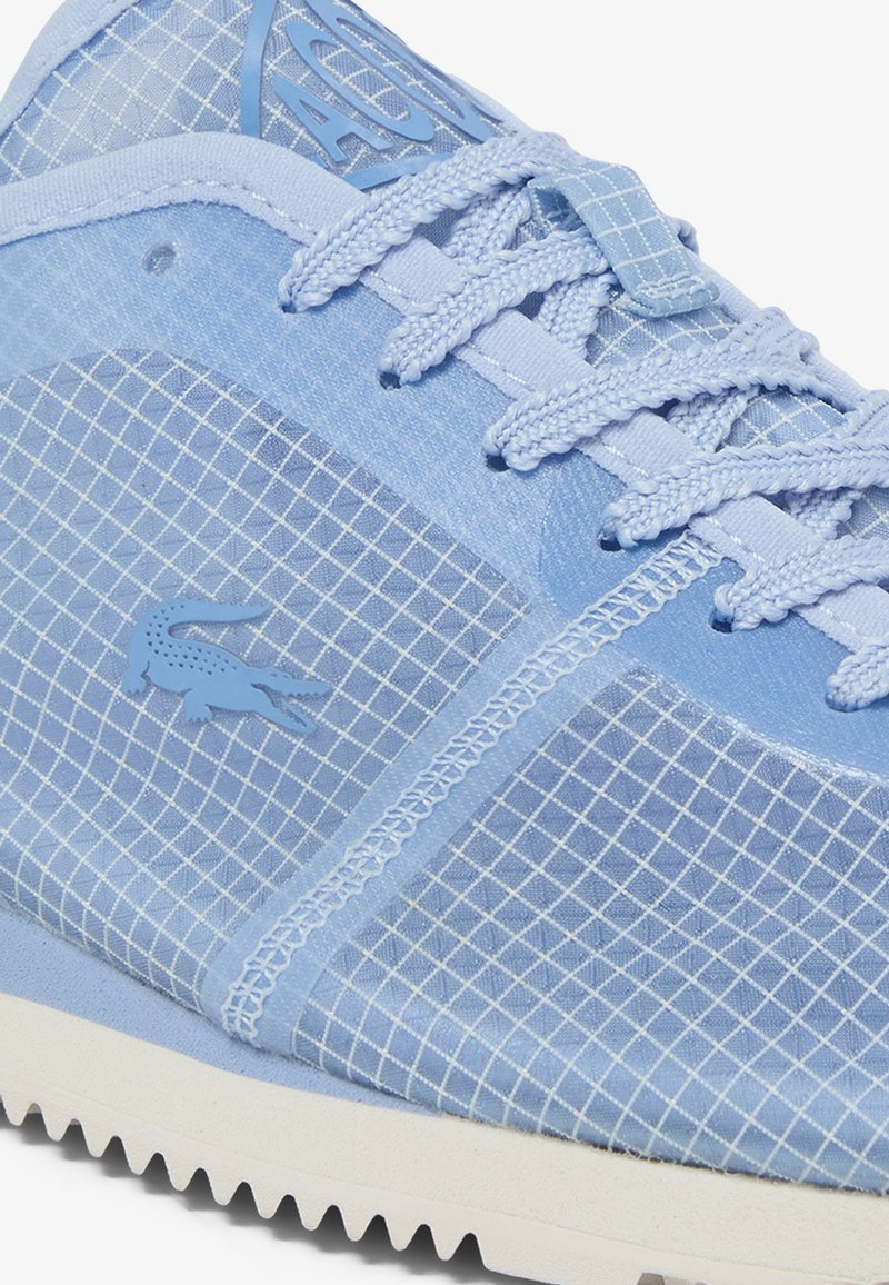 Hellblauer Sneaker mit Rastermuster, strukturierten Schnürsenkeln, abgesteppten Paneelen, Krokodillogo und weißer, gerillter Sohle.