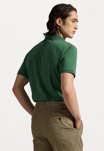 Zaļš polo krekls ar gludu tekstūru, īsām piedurknēm un klasisku apkakli, saskaņots ar brūniem raibiem biksēm, kurām ir aizmugurējā kabata.