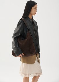 Junge Frau in einer schwarzen Lederjacke, beiger Rock mit Spitzenrand, die eine große braune Wildleder-Schultertasche hält und nach rechts blickt.
