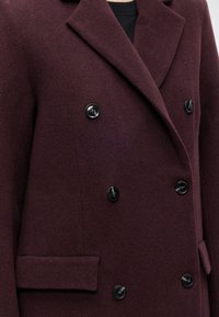 Manteau en laine bordeaux foncé à double boutonnage avec des boutons noirs, larges revers et poches à rabat, porté sur un top noir.
