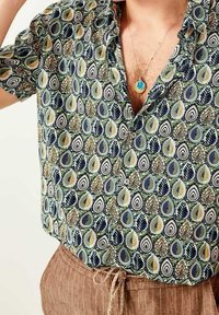 Kurzarmhemd aus grünem, gemustertem Stoff mit tropfen- und blattförmigen Designs in Blau, Gelb und Schwarz. Sichtbare braune, strukturierte Hose.