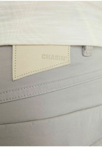 Béžové kalhoty s vyšitým koženým štítkem označeným "CHASIN'" nošené s zasunutou světle béžovou košilí, která je viditelná v pase.