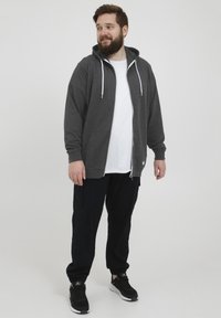 Sudadera gris con cremallera y cordones blancos, con un bolsillo tipo canguro, llevada sobre una camiseta blanca y combinada con pantalones jogger negros y zapatillas.