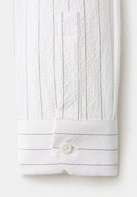 Manche de chemise blanche avec fines rayures noires verticales, un seul bouton blanc sur le poignet, tissu texturé visible.