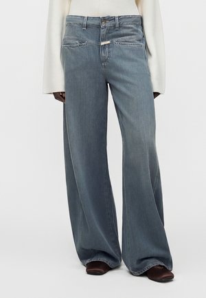 Højttaljede vidde jeans med totonet blå denim, frontlommer, knaplukning, båret med brune sko med firkantet tå og en hvid top med lange ærmer.