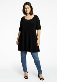 Tunique noire à manches courtes avec un col rond, une forme fluide et un tissu doux, associée à un jean bleu ajusté et des talons noirs.