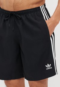 Sorte svømmebukser laget av lett materiale. Har hvite side-striper og en brodert Adidas-logo på venstre ben. Elastisk livlinje.
