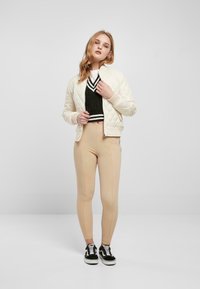 Quilting krem bomberjakke, svart kort genser, beige leggings, og svarte joggesko med hvite snører, satt mot en lys bakgrunn.