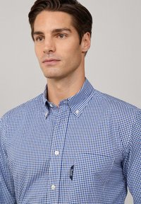 Man die een blauw-wit geruit overhemd met knoopsluiting, een borstzak en een dichtgeknoopte kraag draagt, iets naar links kijkt tegen een effen achtergrond.
