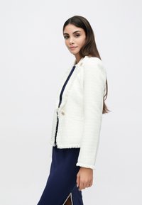 Blazer en tweed blanc avec bords effilochés, fermeture à un bouton et deux poches, associé à une robe marine avec une fente latérale.