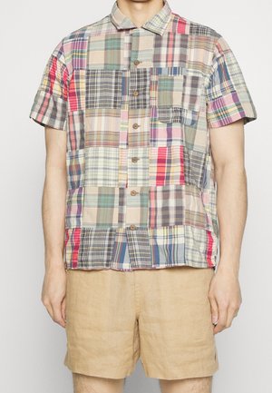 Mand iført kortærmet skjorte med flerfarvet patchworkternet mønster og beige shorts mod hvid baggrund.
