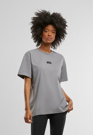 T-shirt in cotone grigio con scollatura a girocollo, maniche corte e un'etichetta nera sul davanti. Fit semplice e rilassato, abbinato a pantaloni neri.