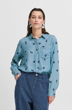 Camicia azzurra chiara in morbido tessuto con ricami floreali blu scuro, taschino frontale e chiusura a bottoni, abbinata a pantaloni a strisce blu scuro testurizzati.
