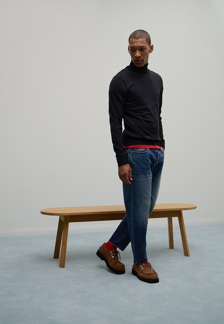 Mężczyzna w czarnym golfie, niebieskich dżinsach, czerwonych skarpetach i brązowych butach stoi obok drewnianej ławki na jasnoniebieskiej podłodze.