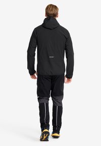 Svart vattentät jacka med huva, lättviktstyg, ergonomisk passform och reflexdetaljer. Kombinerad med svarta och grå byxor samt gula skor.