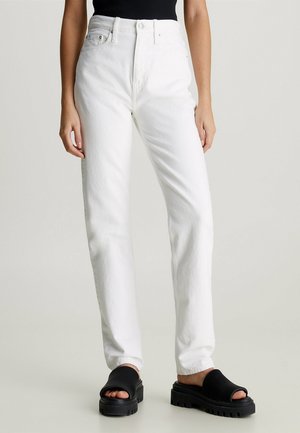 Kvinde iført hvide, højtaljede, lige jeans med forlommer og sorte, chunky slidesandaler, stående mod en ensfarvet baggrund.