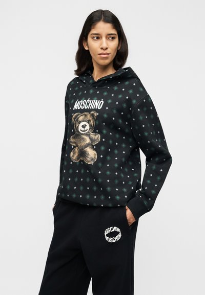 Μαύρο φούτερ με κουκούλα με γραφικό αρκουδάκι και κείμενο "MOSCHINO", με σχέδιο από πράσινα διαμάντια. Συνδυάζεται με μαύρη φόρμα, λογότυπο στην τσέπη.