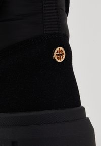 Încălțăminte neagră cu un superior din suede texturat și o talpă din cauciuc negru. Detaliu notabil: un buton decorativ auriu cu un design floral.